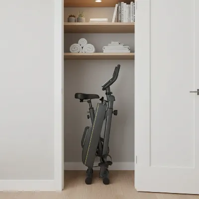 A stylish folding exercise bike in its fully collapsed state, stored upright in a narrow closet space in an apartment, demonstrating its compact storage capability. The bike is dark grey with yellow accents.